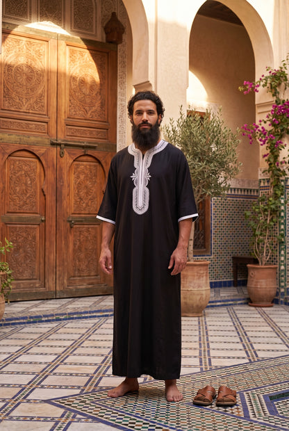 A man wearing a black traditional long Morrocan thobe with white embroidery on the chest stands barefoot in a Moroccan courtyard with patterned tile floors, wooden doors, and potted plants.