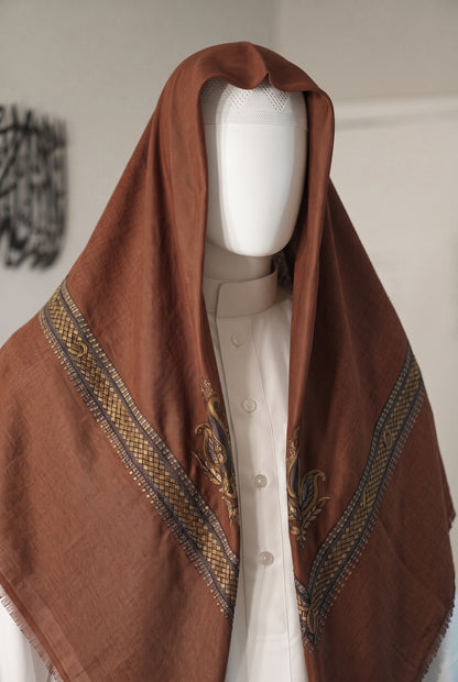 A brown shemagh with intricate gold and blue patterned borders and embroidery, draped over a white mannequin torso wearing a cream thobe.
