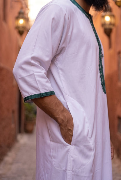 A man wearing a white traditional long Morrocan thobe with dark green embroidered trim on the collar and cuffs, standing in a warm-toned outdoor alleyway.