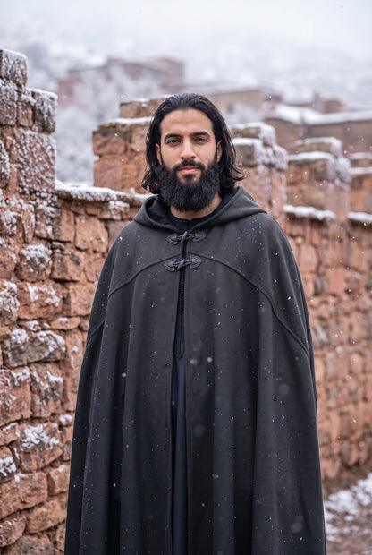 A man with a beard and long dark hair stands outdoors wearing a long black hooded cloak with toggle closures. Snow is lightly falling, and he is posed in front of rustic reddish-brown stone walls with snow-dusted buildings in the background.Arabian Burnous – Hooded Winter Cloak Cape with Toggle Closure | Warm Oversized Djellaba Coat for Men