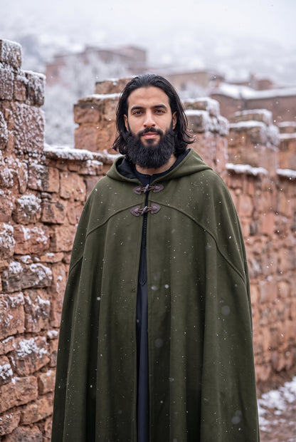 A man with a beard and long dark hair stands outdoors in front of snow-dusted stone walls, wearing an olive green medieval-style hooded cloak with toggle closures over a black garment.Arabian Burnous – Hooded Winter Cloak Cape with Toggle Closure | Warm Oversized Djellaba Coat for Men