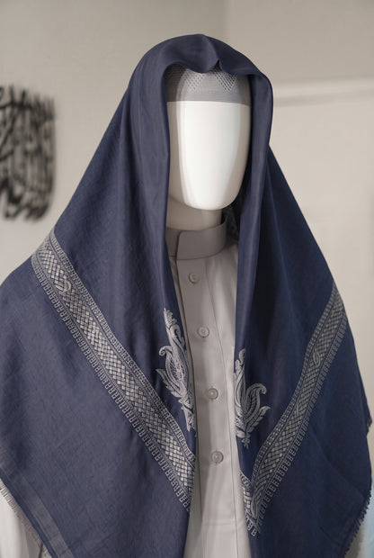 A navy blue shemagh with intricate silver-gray embroidered borders and motifs, draped over a white mannequin torso wearing a light gray button-up shirt.
