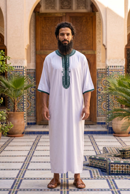 A man with a beard wearing a white traditional Moroccan Morrocan thobe with green embroidery on the chest and sleeve cuffs, standing in a courtyard with tiled floors and wooden door