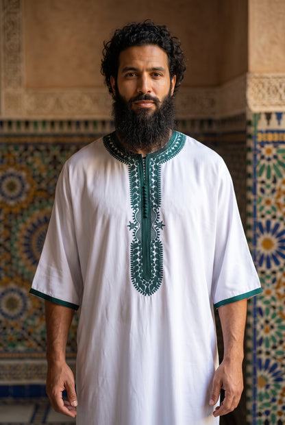 A bearded man with curly hair wearing a white traditional Morrocan thobe with green embroidery on the chest and sleeve cuffs, standing in front of a patterned tile wall.