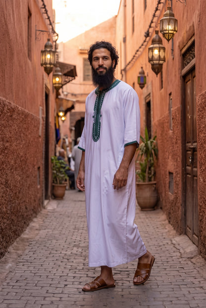 A man wearing a white traditional long Morrocan thobe with green embroidery on the chest and collar, walking down a cobblestone alleyway with warm-toned walls and hanging lanterns.