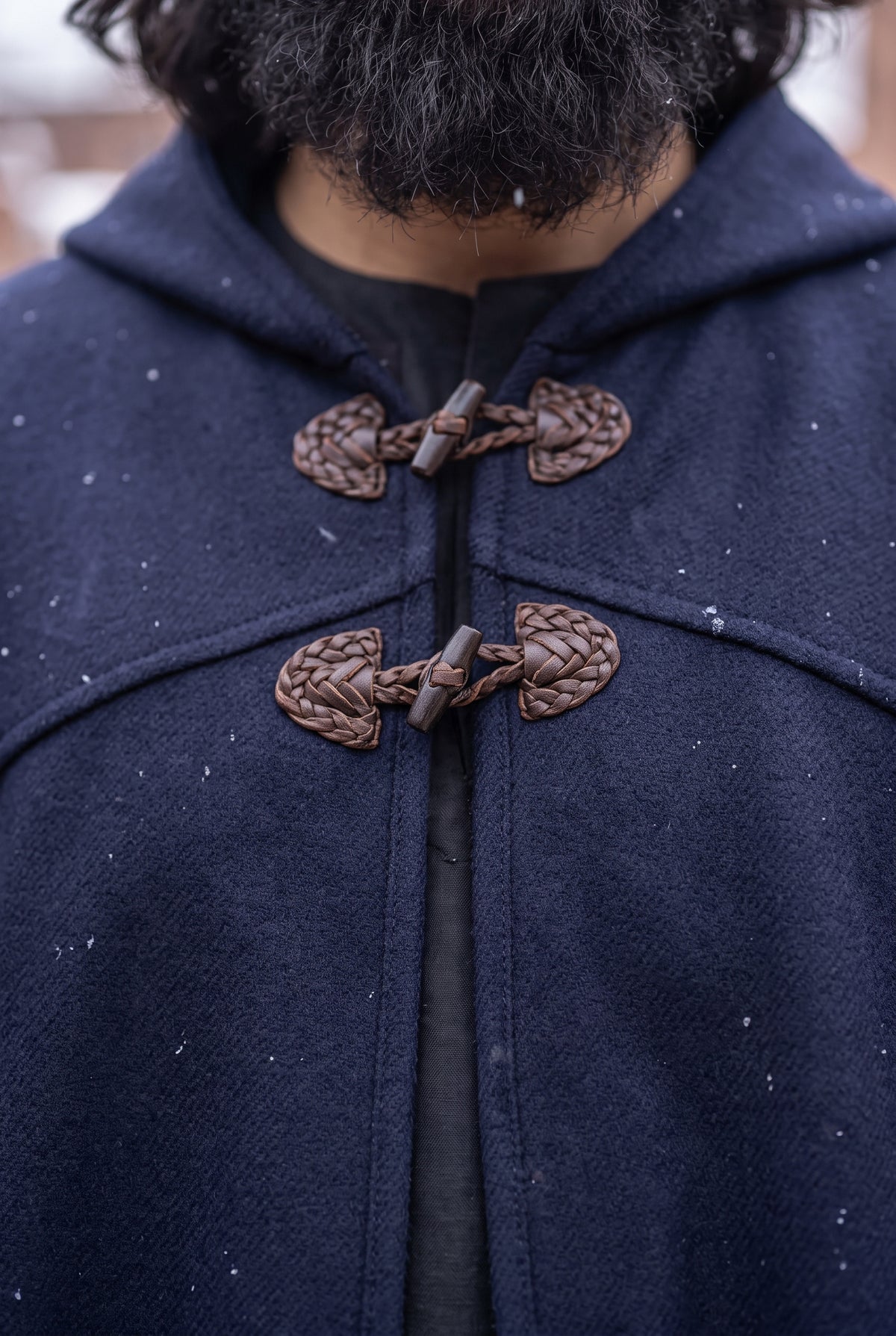 Close-up of a person wearing a dark navy hooded cape or duffle coat with braided brown toggle closures over a black shirt, with snow visible in the background.Arabian Burnous – Hooded Winter Cloak Cape with Toggle Closure | Warm Oversized Djellaba Coat for Men