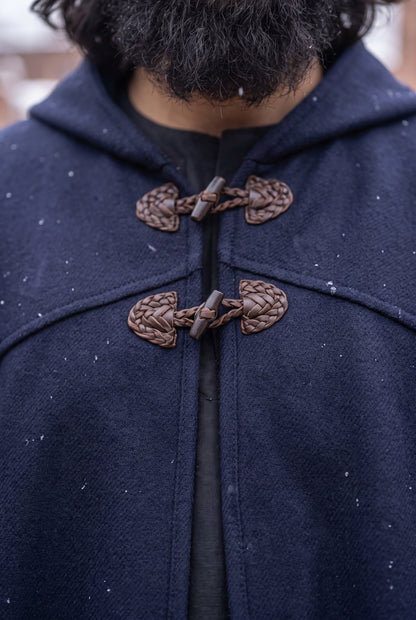 Close-up of a person wearing a dark navy hooded cape or duffle coat with braided brown toggle closures over a black shirt, with snow visible in the background.Arabian Burnous – Hooded Winter Cloak Cape with Toggle Closure | Warm Oversized Djellaba Coat for Men