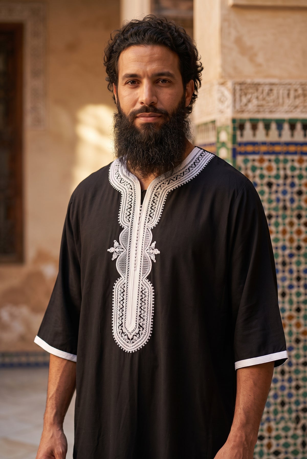 A bearded man wearing a black traditional tunic with white Morrocan thobe embroidered detailing on the chest and sleeve cuffs, standing in front of a tiled architectural wall.