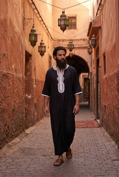 A man with a beard wearing a black traditional Moroccan Morrocan thobe with white embroidery walks down a narrow cobblestone alleyway with warm, earth-toned walls and hanging lanterns.