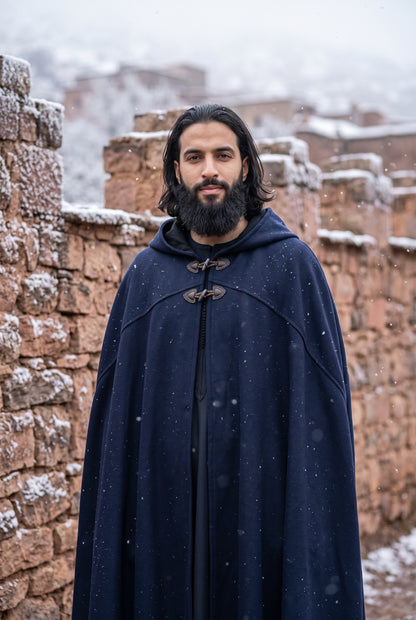 A man with a beard and long dark hair stands outdoors in a snowy setting, wearing a long navy blue hooded cloak with toggle closures. The background shows rustic stone walls with snow on top.Arabian Burnous – Hooded Winter Cloak Cape with Toggle Closure | Warm Oversized Djellaba Coat for Men