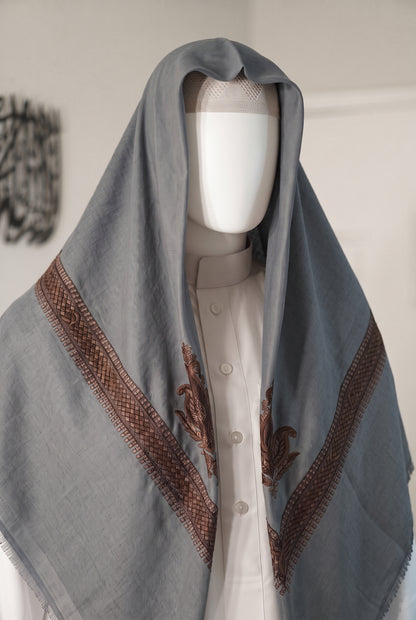 A slate blue-gray shemagh with brown embroidered borders and motifs, draped over a white mannequin torso wearing a white thobe.