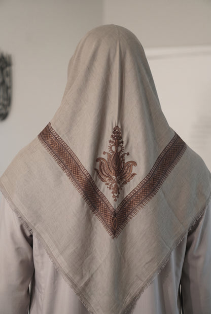 A light beige linen shemagh with brown embroidered borders and a central floral motif, draped over the head of a person shown from behind.